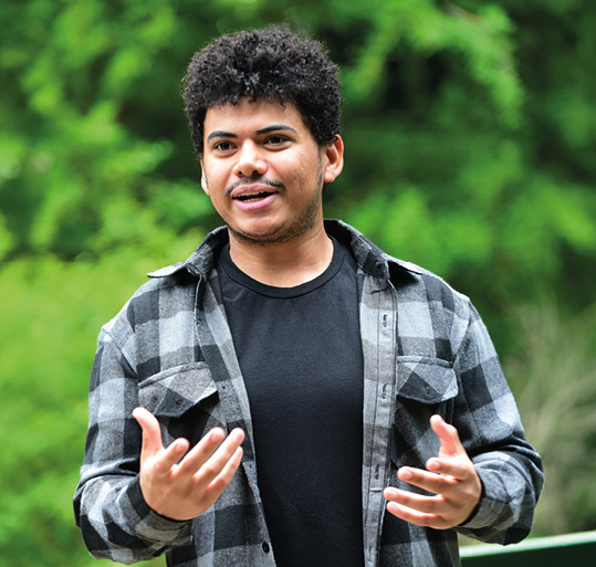 Jovem com camisa xadrez cinza e preta, gesticulando com as mãos em um ambiente externo com fundo verde desfocado.