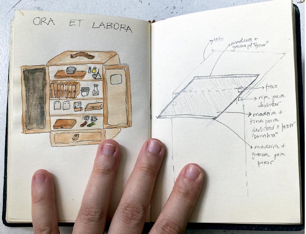 Aqui está a descrição da imagem focada em acessibilidade:

A imagem mostra um caderno de esboços (sketchbook) aberto, apoiado em uma superfície clara. Na parte inferior, vê-se os dedos de uma mão de pele clara pressionando as páginas para mantê-las abertas.

Página da Esquerda: No topo, está escrita a frase em latim "ORA ET LABORA" (que significa "ore e trabalhe") em letras maiúsculas manuscritas. Abaixo da frase, há um desenho colorido, feito com traços soltos (estilo aquarela ou marcador), representando um armário de madeira antigo. O armário tem duas portas abertas, revelando prateleiras cheias de pequenos objetos indefinidos (que parecem ser livros, frascos e caixas) e uma gaveta na base.

Página da Direita: Esta página contém um esboço técnico feito a lápis grafite. O desenho é um diagrama tridimensional de uma estrutura retangular (semelhante a uma tampa ou caixa), com várias setas e anotações manuscritas em português ao redor. As anotações descrevem detalhes construtivos, com termos legíveis como "teto", "furo", "ripa para deslizar", "madeira + fina para deslizar e fazer 'sombra'" e "madeira + grossa para fixar".