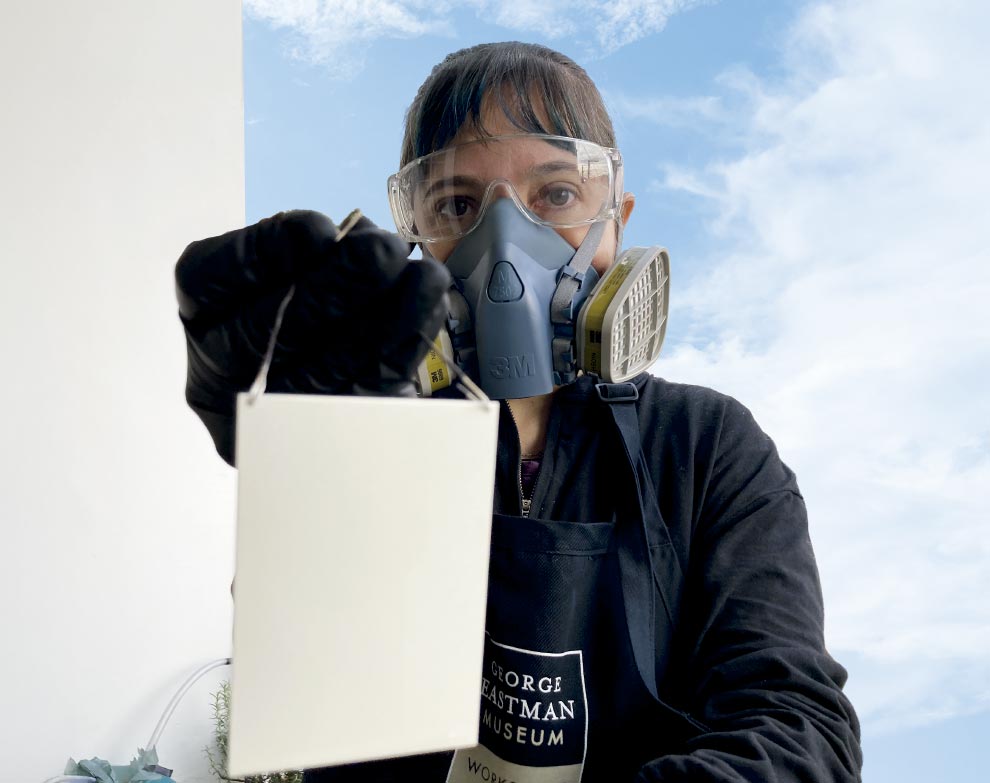 A imagem mostra uma pessoa (aparentemente uma mulher de pele clara) em plano médio, olhando diretamente para a câmera. Ela está equipada com proteção de segurança industrial: usa óculos de proteção transparentes e uma máscara respiratória grande (tipo respirador semifacial) nas cores cinza e azul, que cobre o nariz e a boca, com filtros nas laterais.

Seu cabelo é escuro, com franja e algumas mechas tingidas de azul. Ela veste uma camisa preta de manga comprida e um avental preto onde se lê, em letras brancas dentro de um quadrado: "GEORGE EASTMAN MUSEUM".

Com a mão direita, coberta por uma luva preta de proteção, ela segura e estende em direção à câmera uma pequena placa retangular branca lisa, que está pendurada por uma alça de arame fino. O gesto coloca o objeto em destaque no primeiro plano.

O fundo é um céu azul claro com nuvens brancas dispersas, sugerindo um ambiente externo. À esquerda, vê-se um fragmento de uma parede branca. A iluminação é natural e clara.