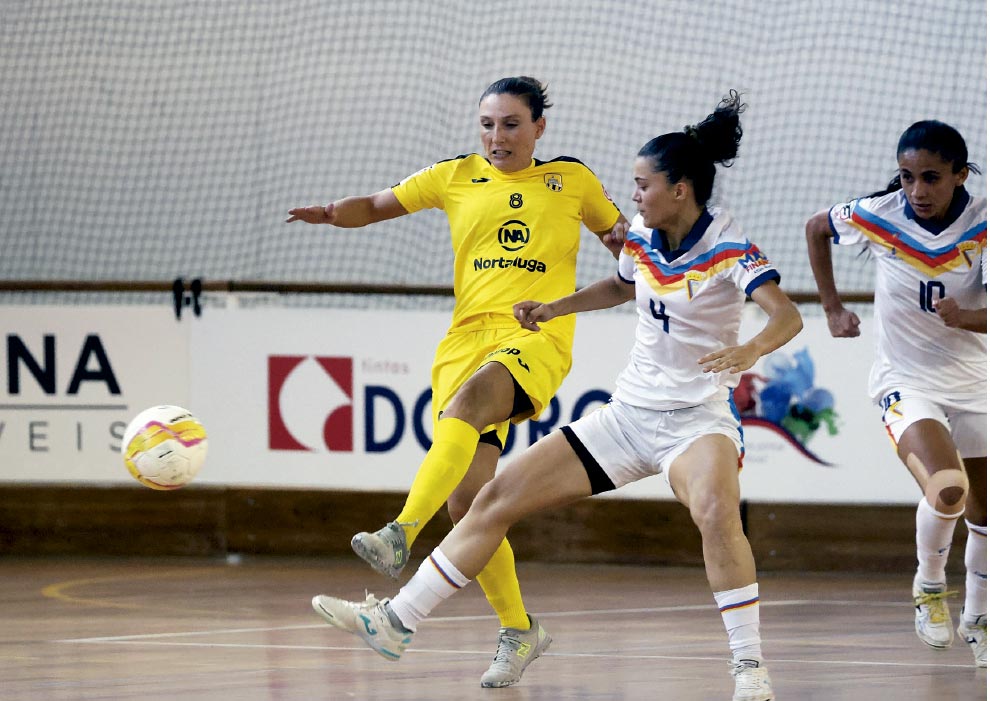 A imagem captura um momento dinâmico de uma partida de futsal feminino em uma quadra coberta. A cena foca na disputa de bola entre três jogadoras.

No centro, uma jogadora vestindo uniforme completo amarelo (camisa número 8) está com a perna esquerda levantada no ar, acabando de chutar a bola. A bola, que é branca com detalhes em amarelo e rosa, está suspensa no ar, próxima ao pé dela.

Tentando bloquear a jogada, logo à frente e um pouco à direita, está uma adversária de uniforme branco com uma faixa em formato de "V" no peito nas cores amarelo, azul e vermelho (o número 4 está visível na camisa). Ela está com as pernas afastadas em posição de defesa e olhar focado na bola.

No canto direito da imagem, uma terceira jogadora, companheira da que está defendendo (mesmo uniforme branco, número 10), corre em direção ao lance.

O piso da quadra é de madeira polida. Ao fundo, fora de foco, há placas de publicidade brancas com logotipos e textos (como "Tintas") e uma rede de proteção branca cobrindo a parede.