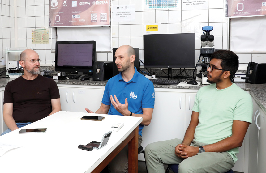 Três homens estão sentados à mesa em um laboratório. Um deles está falando e gesticulando.