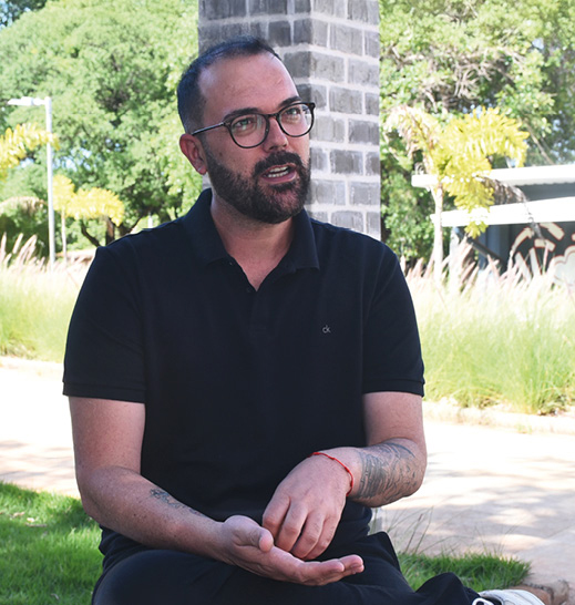 Homem de barba, usando óculos e camisa polo preta. Ele está sentado ao ar livre, próximo a uma coluna de tijolos, com árvores e grama no fundo.