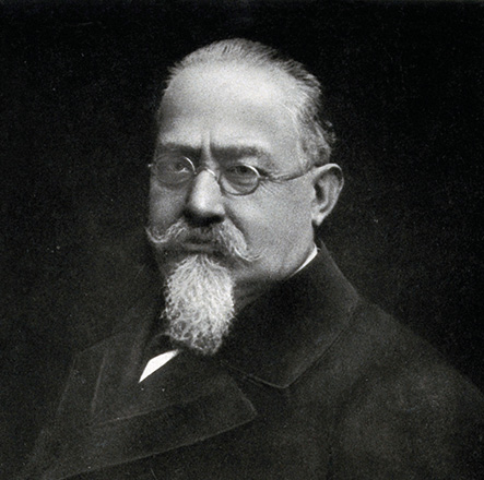 É uma foto antiga em preto e branco de um homem de meia-idade, com óculos e um grande bigode. Ele tem uma barba longa e pontuda (cavanhaque) e veste um casaco escuro. O fundo é preto.