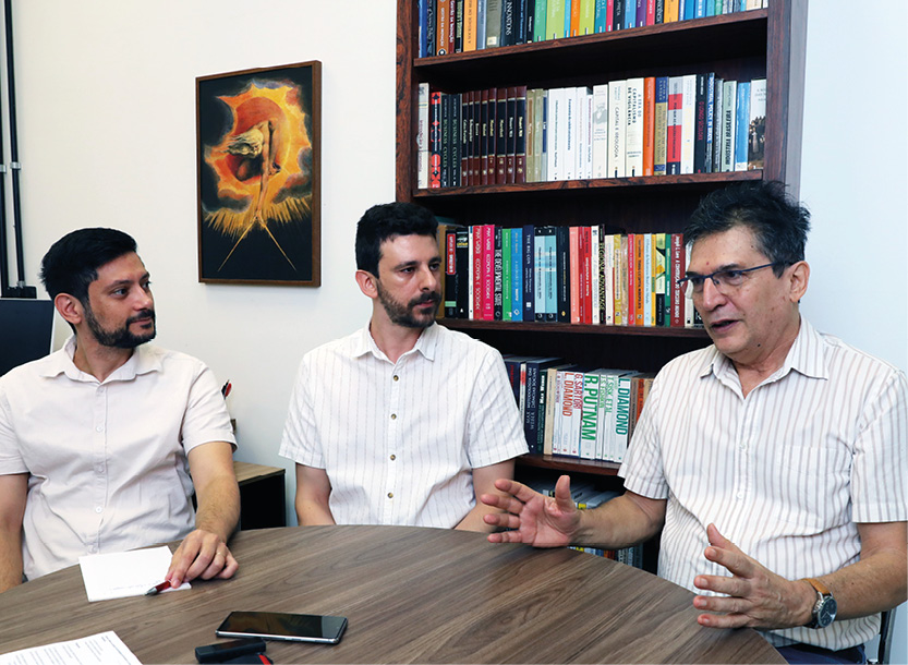 Três homens estão sentados ao redor de uma mesa redonda de madeira em um escritório. O homem à direita, de camisa listrada e óculos, gesticula com as mãos enquanto fala, sendo observado pelos outros dois, que vestem camisas claras de manga curta. Ao fundo, há uma estante repleta de livros e um quadro na parede branca.