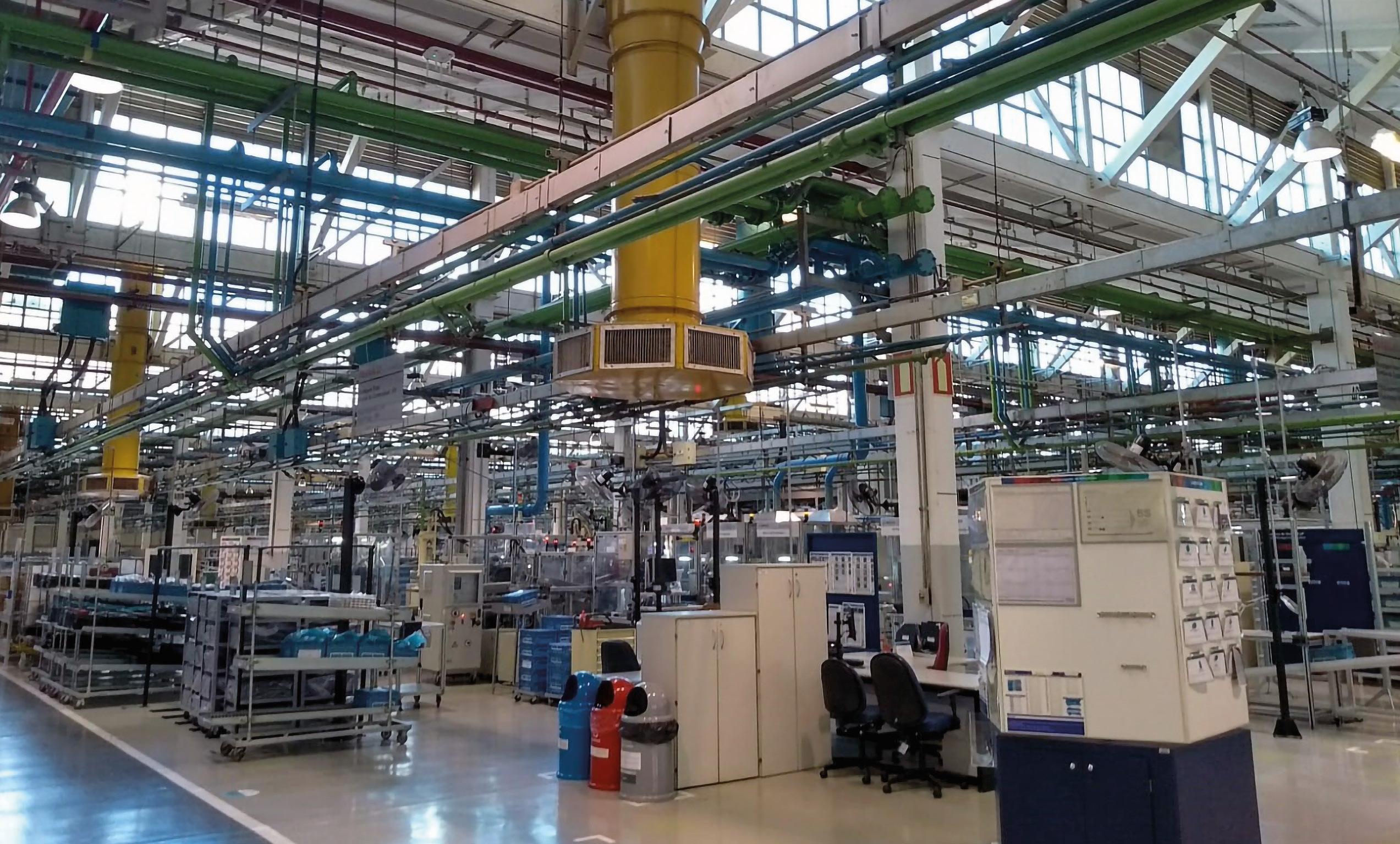 Uma ampla área interna de uma fábrica, com o teto alto tomado por tubulações industriais coloridas em verde, azul e amarelo. No chão, veem-se bancadas de trabalho, carrinhos de transporte, lixeiras coloridas de coleta seletiva e uma estação de escritório com cadeiras e computadores.