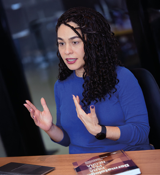 A imagem mostra uma mulher com cabelos escuros e longos, combinando tranças finas no topo e cachos definidos, vestindo uma blusa de manga comprida azul royal. Ela está sentada à frente de uma mesa de madeira, com as mãos levantadas em um gesto explicativo enquanto fala.
