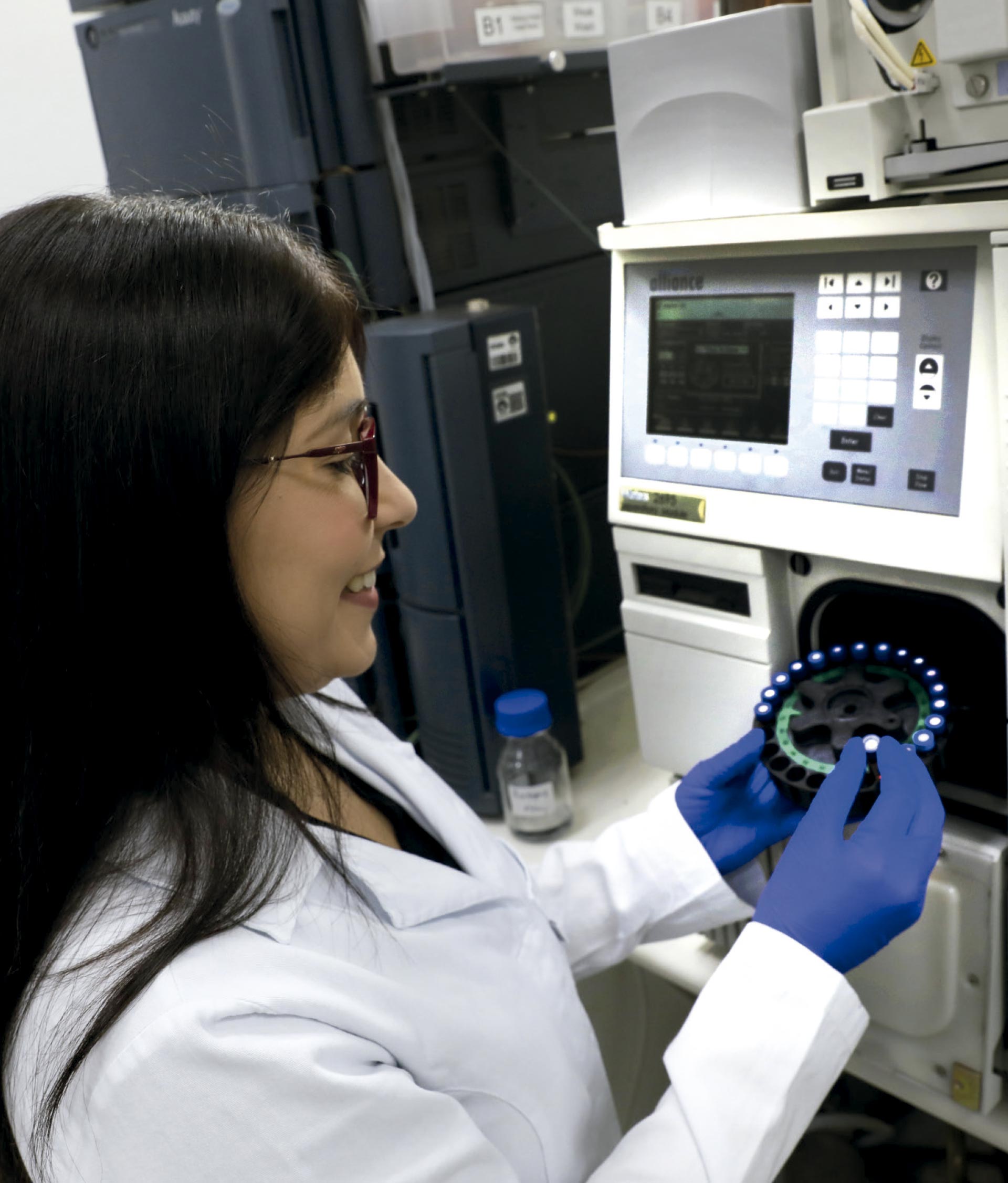 Uma mulher em um laboratório aparece de perfil, sorrindo enquanto trabalha em um equipamento científico. Ela usa jaleco branco, óculos e luvas azuis, e segura com as duas mãos uma peça circular com vários pequenos tubos iluminados ao redor. À sua frente há uma máquina grande com tela e botões, onde ela parece encaixar ou ajustar a peça. Ao fundo, aparecem outros equipamentos de laboratório e um pequeno frasco sobre a bancada, indicando um ambiente de pesquisa ou análise científica.